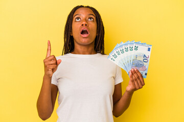 Young african American woman holding bills isolated on yellow background  pointing upside with opened mouth.