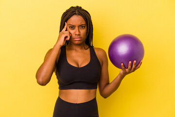 Young african american woman holding a pilates ball isolated on yellow background  pointing temple with finger, thinking, focused on a task.