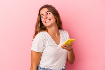 Young caucasian woman using mobile phone isolated on pink background  looks aside smiling, cheerful and pleasant.