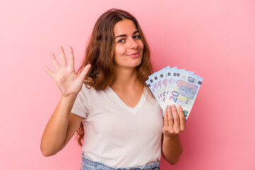 Young caucasian woman holding bills isolated on pink background  smiling cheerful showing number five with fingers.