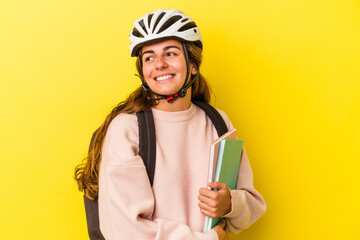 Young caucasian student woman wearing a bike helmet isolated on yellow background  looks aside smiling, cheerful and pleasant.