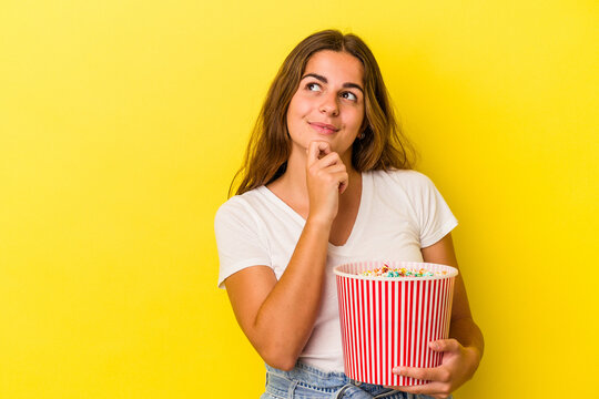 Young caucasian woman holding a popcorns isolated on yellow background  looking sideways with doubtful and skeptical expression.