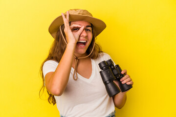 Young caucasian woman holding binoculars isolated on yellow background  excited keeping ok gesture on eye.