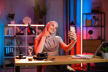 Fototapeta na wymiar Pretty muslim woman taking selfie on smartphone while sitting at home office. Wireless laptop on table. Dark atmosphere at room. Concept of gadgets and freelance.