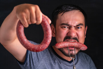 A bearded, hungry man greedily eats a huge piece of sausage impaled on a fork. holding a whole circle of sausage in the other.