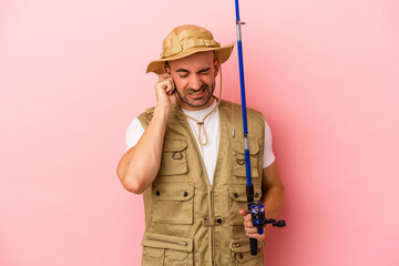 Young bald fisherman holding a rod isolated on pink background  covering ears with hands.