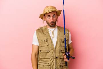 Young bald fisherman holding a rod isolated on pink background  shrugs shoulders and open eyes confused.