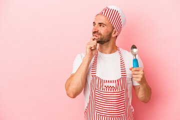 Young ice cream maker man holding spoon isolated on pink background  looking sideways with doubtful and skeptical expression.