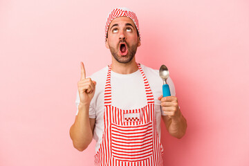 Young ice cream maker man holding spoon isolated on pink background  pointing upside with opened mouth.