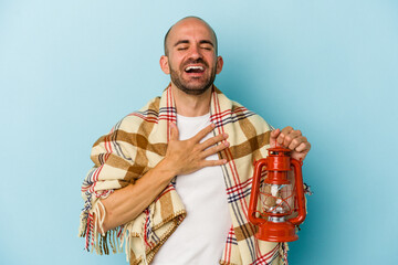 Young bald man holding vintage lantern isolated on blue background  laughs out loudly keeping hand on chest.