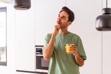 Young mixed race man drinking orange juice in his kitchen looking sideways with doubtful and...