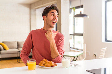 Fototapeta na wymiar Young mixed race man having breakfast in a kitchen on the morning points with thumb finger away, laughing and carefree.
