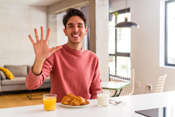 Fototapeta na wymiar Young mixed race man having breakfast in a kitchen on the morning smiling cheerful showing number five with fingers.
