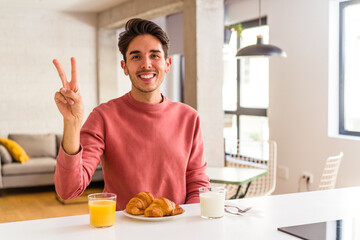 Fototapeta na wymiar Young mixed race man having breakfast in a kitchen on the morning joyful and carefree showing a peace symbol with fingers.