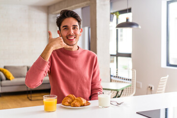 Fototapeta na wymiar Young mixed race man having breakfast in a kitchen on the morning showing a mobile phone call gesture with fingers.