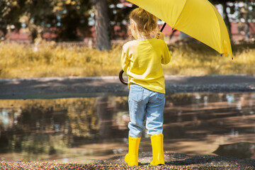 A child, a girl in yellow clothes and rubber boots with an umbrella stands near a puddle on an autumn walk.