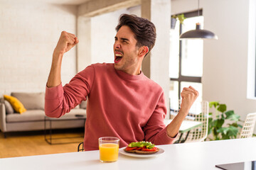 Fototapeta na wymiar Young mixed race man having breakfast in his kitchen raising fist after a victory, winner concept.