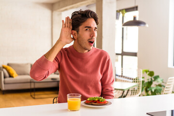 Young mixed race man having breakfast in his kitchen trying to listening a gossip.