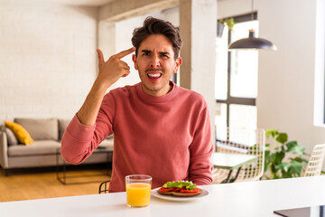 Fototapeta na wymiar Young mixed race man having breakfast in his kitchen showing a disappointment gesture with forefinger.