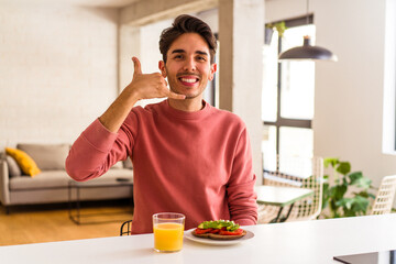Fototapeta na wymiar Young mixed race man having breakfast in his kitchen showing a mobile phone call gesture with fingers.