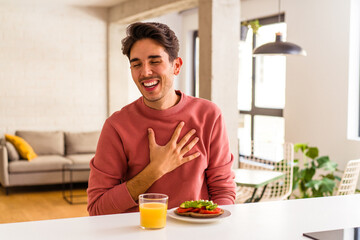Fototapeta na wymiar Young mixed race man having breakfast in his kitchen laughs out loudly keeping hand on chest.