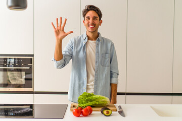 Young mixed race man preparing a salad for lunch smiling cheerful showing number five with fingers.