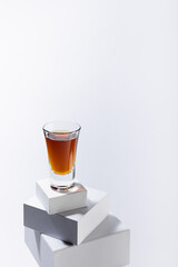 Brown old cognac in shot glass on stack of white boxes as podium in soft light white abstract space, luxury minimal geometric style, vertical.