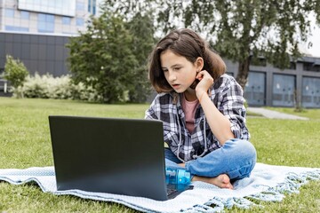 A schoolgirl watches an Internet class and studies remotely. The girl uses wireless internet technologies. A pretty young teenage girl sitting on the grass and working on a laptop.