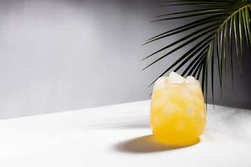 Misted tropical orange juice with ice cubes in glass on podium in sunlight with shadows, palm leaf in white and dark black abstract space or outdoor.