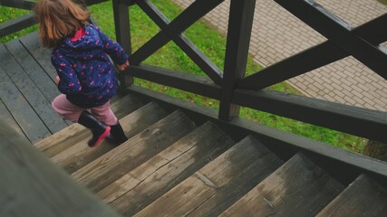 Caucasian Girl Running Down Cabin External Wooden Stairs Wearing Rain Jacket and Gum Welligtons...