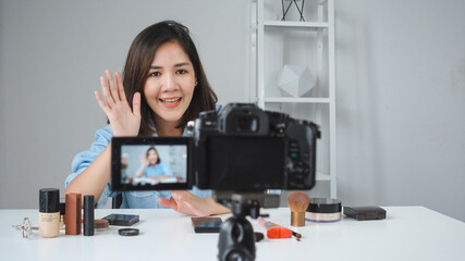 Happy smiling asian woman or beauty blogger with brush and camera recording video and waving hand at home. Beauty videoblog blogging people concept.