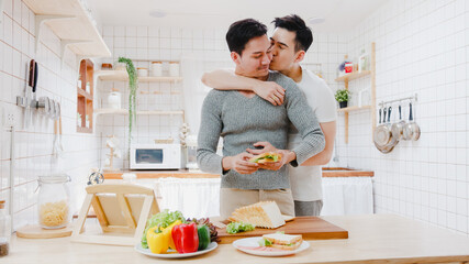 Asia gay couple using tablet and preparing the breakfast, sandwich vegetable on table in kitchen at...