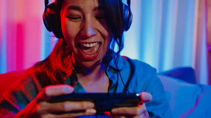 Happy asia girl gamer wear headphone competition video game online with smartphone excited talk with friend sit on couch in colorful neon lights living room at home, Home quarantine activity concept.