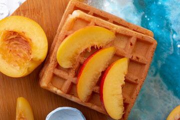 Belgian homemade waffles with peaches and yellow flowers on a beautiful wooden board with a blue pattern. top view. photo for the menu. dessert for breakfast on a white stone background. flat lay
