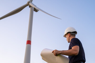 An engineer of wind turbines holding a paper project and looking at windmill in the field. Green eco electricity generation and renewable energy. 