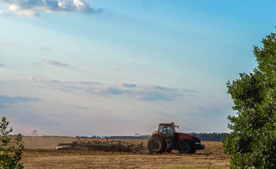 Fototapeta premium The tractor plows the field against the background of the forest. Selective focus.
