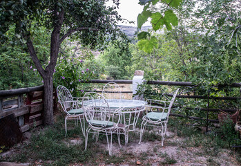 table and four white wrought-iron chairs on a terrace overlooking the countryside with many trees and vegetation next to a thicket with lilac flowers