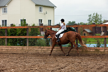 A girl in white equestrian clothes and a helmet, riding a brown horse. Riding a horse.