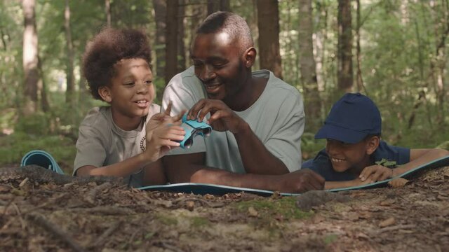 Medium close-up of African American man and his playful little sons having camping trip together, lying on their stomachs on forest ground, looking at animals and birds through binoculars