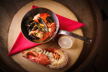 Hot, freshly broiled halved Maine Lobster with lemon and cioppino or bouillabaisse mussels seafood stew soup in pan on wood board and red napkin over wooden background. Rustic setting.Warm tone light.