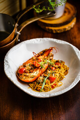 Hot and freshly cooked halved Maine Lobster with Italian Spaghetti pasta with tarragon and tomato on white ceramic plate over wood table. Pan and rustic kitchen setting in background. Warm tone light.