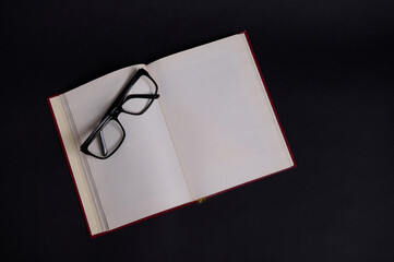 Flat lay of eyeglasses on an opened book in hard red cover, isolated over black background with...
