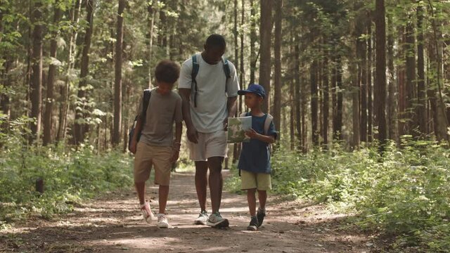 Slowmo full-length shot of African American man and his two little sons walking on forest path on warm sunny day camping or hiking together with backpacks and map