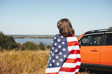 Young woman with American flag standing next to a car in nature. Back view