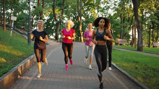 Fitness class for seniors. Group of active mature ladies with young black trainer running in urban park, tracking shot