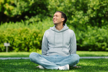 Happy young caucasian man in casual clothing sitting on grass in city park resting during a break from work, relaxed, breathes fresh air, closed eyes, crossed legs.