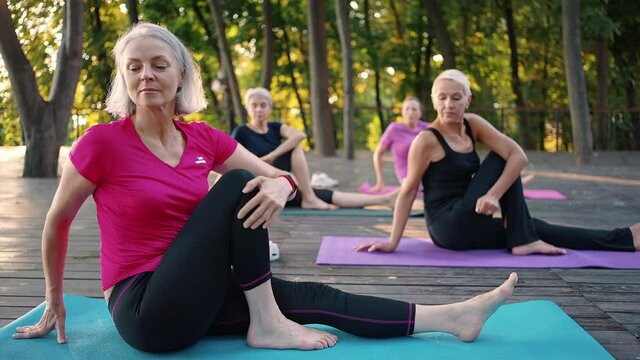 Group of active mature woman training in park, making stretching exercises and yoga asana, sitting on fitness mats