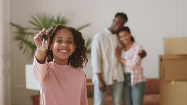 Mortgage for young families. Happy african american little girl showing apartment keys, parents embracing on background