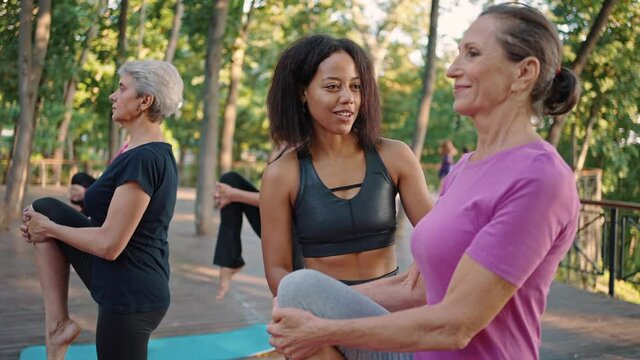 Young black positive female coach correcting senior lady yoga beginner during group class training outdoors