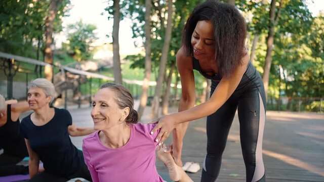 Female yoga instructor correcting class member during group training outdoors, helping senior lady with difficult asana